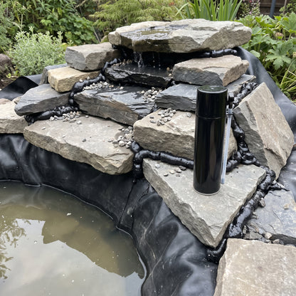 Black waterfall foam sealing the gaps between stones on a garden pond waterfall under construction, with gravel pressed into the foam