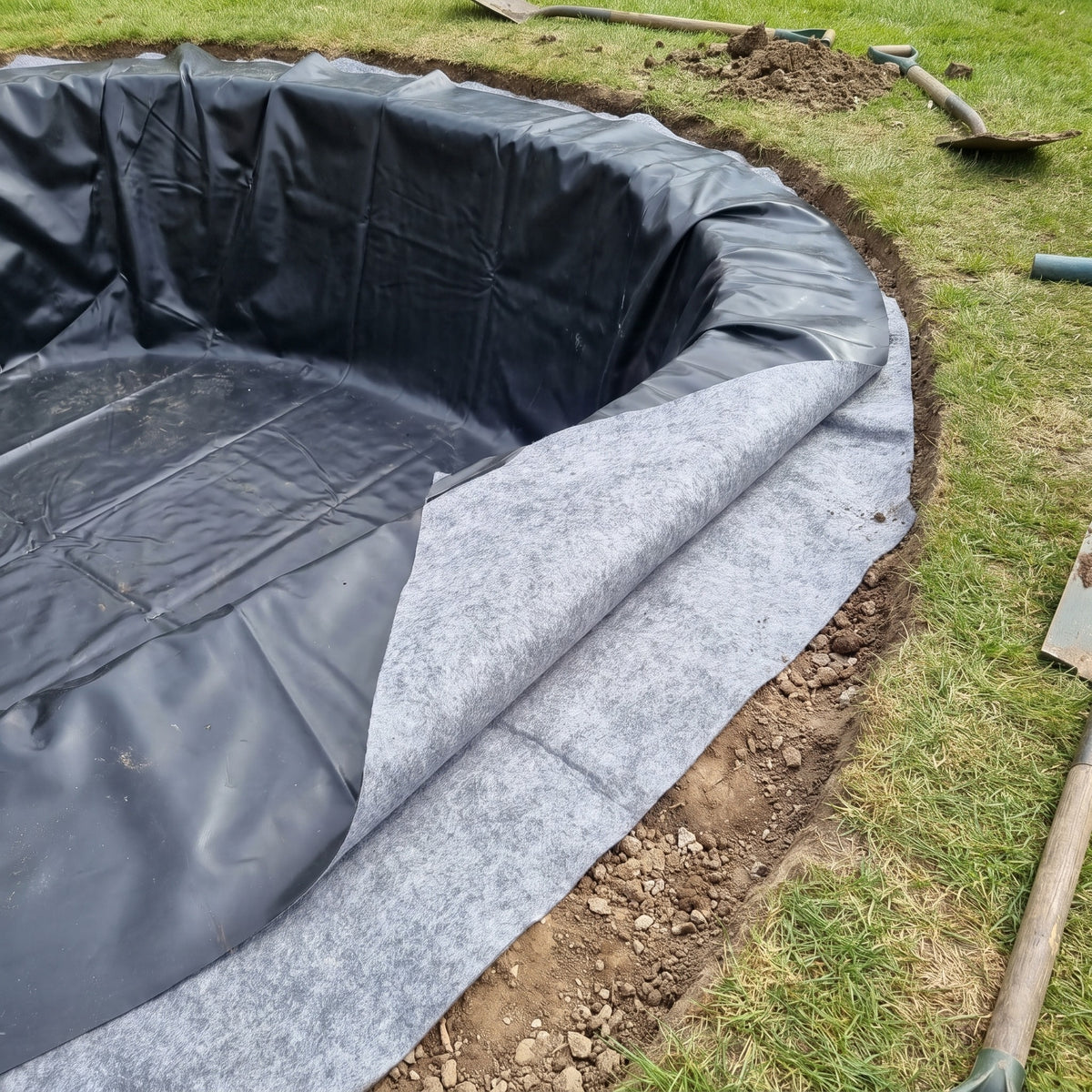 Close-up of black LDPE pond liner laid on top of grey geotextile underlay at the edge of a new pond