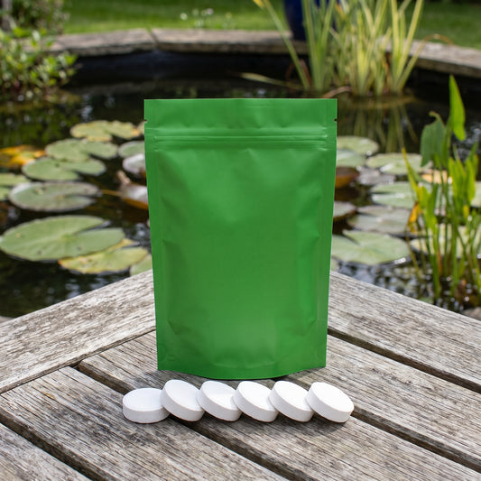 Green pouch of pond sludge reducer tablets with six white tablets lined up on a wooden deck beside a UK lily pond
