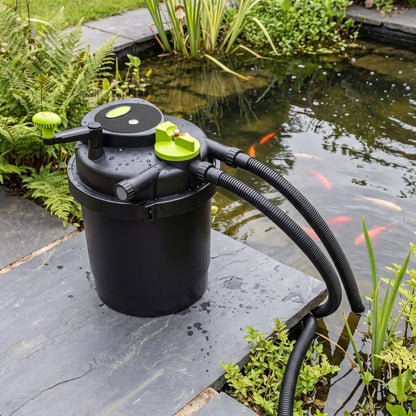 Pressurised UV pond filter with lime-green turn-to-clean handle standing on a slab beside a UK garden fish pond, hoses running into the water