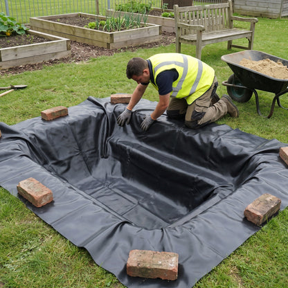 Landscaper pressing a small black PVC pond liner into a freshly dug wildlife pond in a school garden